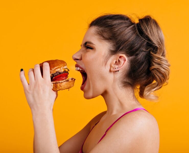 Studio shot of hungry woman with sandwich. Caucasian girl eats hamburger with pleasure