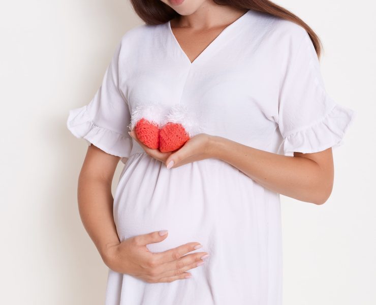 Pregnant happy woman holding red knitted baby shoes in her hands, touching her belly with charming smile, expectant mother wearing white dress, brunette girl looks happy expecting child.